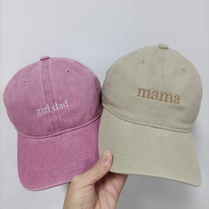 Op de afbeelding: Twee baseballpetten met geborduurde tekst. De linker pet is oudroze met de witte tekst 'girl dad' op de voorkant gestikt. De rechter pet is lichtbeige met de bruine tekst 'mama' op het voorpaneel van de pet geborduurd.