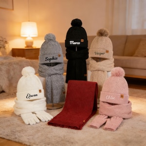 Könnte beinhalten: Eine Sammlung personalisierter Winteraccessoires, darunter Mützen, Schals und Handschuhe. Die Mützen sind in Weiß, Grau, Schwarz und Rosa erhältlich, jede mit einem Namen bestickt. Die Schals sind in Grau, Schwarz, Beige und Rot erhältlich. Die Handschuhe sind in Weiß und Rosa.