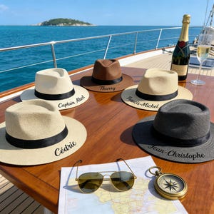 Könnte beinhalten: Sechs Fedora-Hüte in verschiedenen Farben, darunter Weiß, Braun und Schwarz, mit schwarzen Bändern und personalisierten Namen. Die Hüte sind auf einer Holzoberfläche mit Sonnenbrille, Kompass und Karte angeordnet. Eine Flasche Champagner und ein Glas sind ebenfalls vorhanden.