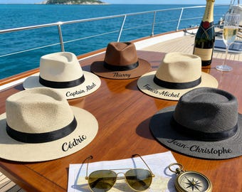 Puede incluir: Seis sombreros fedora en varios colores, incluyendo blanco, marrón y negro, con bandas negras y nombres personalizados. Los sombreros están colocados sobre una superficie de madera con gafas de sol, una brújula y un mapa. También hay una botella de champán y una copa.