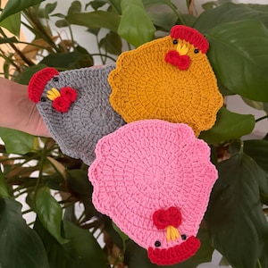Könnte beinhalten: Drei handgefertigte Hühner-Untersetzer aus Häkelarbeit in Grau, Gelb und Rosa. Jeder Untersetzer hat einen roten Kamm und Kehllappen, mit schwarzen Perlenaugen und gelbem Schnabel. Die Untersetzer sind kreisförmig mit einem gewellten Rand.