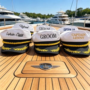 Könnte beinhalten: Mehrere weiße Kapitänsmützen mit schwarzen Krempen und goldenen Akzenten. Einige Mützen tragen Aufschriften wie "Retired" und "Groom". Die Mützen sind vor einem nautischen Emblem auf einer Holzoberfläche angeordnet.