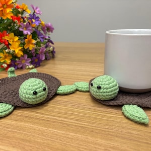 Könnte beinhalten: Zwei handgefertigte Schildkröten-Untersetzer aus Häkelarbeit. Die Untersetzer sind braun mit grünen Flossen und Köpfen. Eine weiße Tasse steht auf einem der Untersetzer. Ein Strauß bunter Blumen befindet sich im Hintergrund.