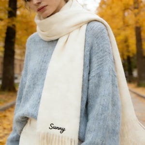 May include: Cream-colored scarf with the word "Sunny" embroidered in black script. The scarf is draped over a light blue sweater. The background features autumn foliage.
