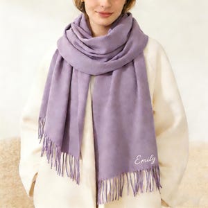 May include: A lavender scarf with fringe detail and the name "Emily" embroidered in white script. The scarf is draped over a white coat. The scarf is a solid color and appears to be made of a soft material.