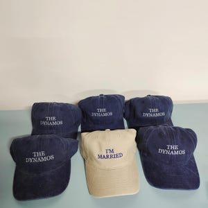 May include: A collection of six baseball caps. Five caps are dark navy blue with the white embroidered text THE DYNAMOS. One cap is beige with the blue embroidered text I'M MARRIED. Each cap features a curved brim and a structured crown design.