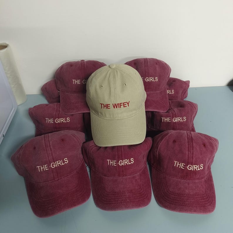 May include: A collection of baseball caps featuring one beige cap embroidered with the red text THE WIFEY and several maroon caps embroidered with the white text THE GIRLS. Each cap has a structured crown, a curved brim, and a button on top.