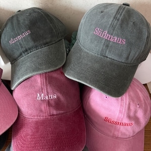 Puede incluir: Una colección de cuatro gorras de béisbol en gris carbón, rosa oscuro y rosa claro. Las gorras gris carbón presentan el texto bordado 'süssmaus' y 'Süßmaus' en rosa. La gorra rosa oscuro tiene 'Maus' en blanco y la rosa claro tiene 'Süßmaus' en rosa.