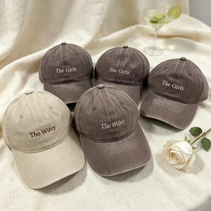 May include: Five baseball caps with a distressed finish. Three dark brown caps are embroidered with the text The Girls in white. One beige cap and one dark brown cap are embroidered with the text The Wifey in dark brown and white respectively.