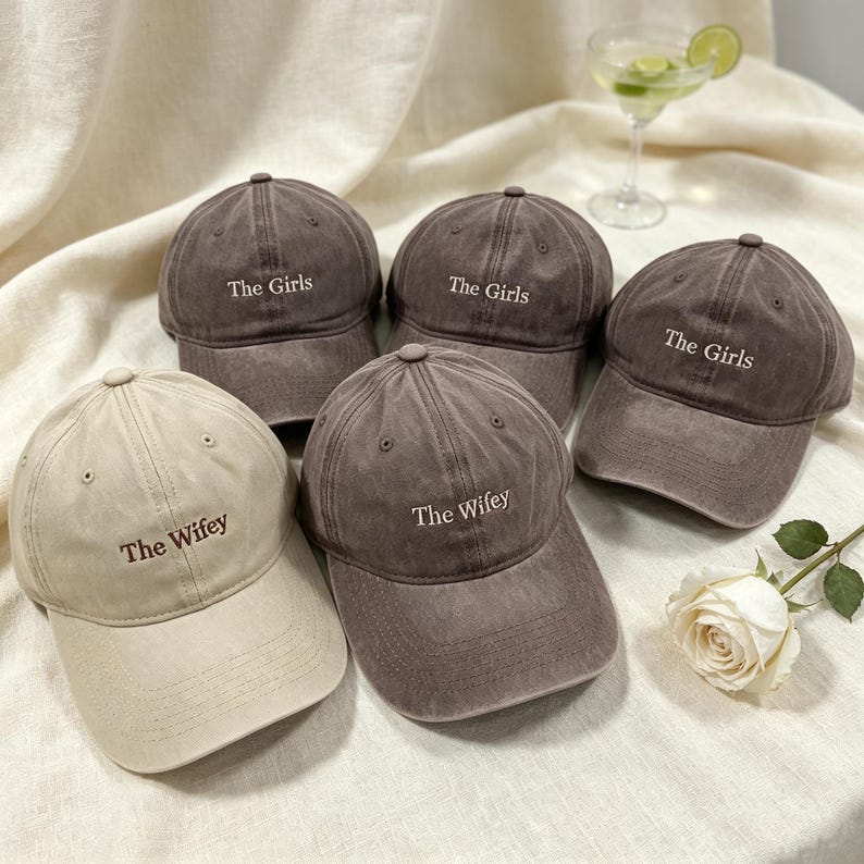 May include: Five baseball caps with a distressed finish. Three dark brown caps are embroidered with the text The Girls in white. One beige cap and one dark brown cap are embroidered with the text The Wifey in dark brown and white respectively.