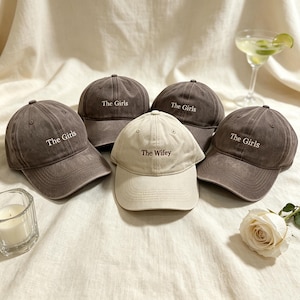 May include: Four dark gray baseball caps embroidered with the white text The Girls and one cream baseball cap embroidered with the brown text The Wifey. Each cap features a curved brim, a six-panel construction, and a button on the top crown.