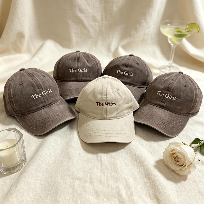 May include: Four dark gray baseball caps embroidered with the white text The Girls and one cream baseball cap embroidered with the brown text The Wifey. Each cap features a curved brim, a six-panel construction, and a button on the top crown.