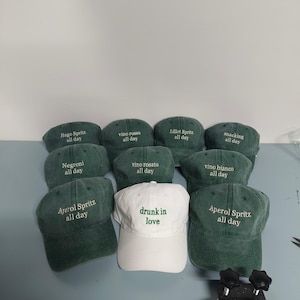 May include: A collection of baseball caps in dark green and white. The green caps feature embroidered text including Hugo Spritz, vino rosso, Lillet Spritz, snacking, Negroni, vino rosato, vino bianco, and Aperol Spritz all day. One white cap says drunk in love.