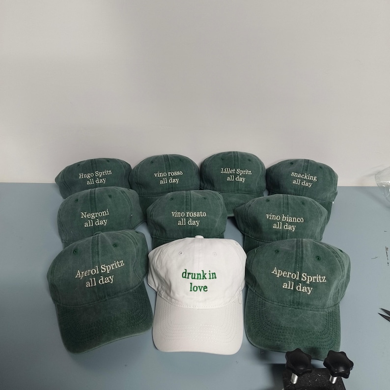 May include: A collection of baseball caps in dark green and white. The green caps feature embroidered text including Hugo Spritz, vino rosso, Lillet Spritz, snacking, Negroni, vino rosato, vino bianco, and Aperol Spritz all day. One white cap says drunk in love.