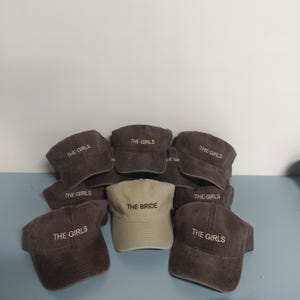 May include: A collection of baseball caps featuring one light beige cap embroidered with the text THE BRIDE and several dark gray caps embroidered with the text THE GIRLS. Each cap has a curved brim and a structured crown design.