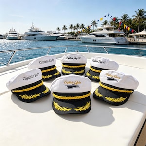 Könnte beinhalten: Sechs weiße Kapitänsmützen mit schwarzen Schirmen und goldenen Verzierungen sind auf einem Bootsdeck angeordnet. Jede Mütze hat einen individuellen Text in Schwarz oder Gold, wie "Captain Ricky" und "Captain Gala". Im Hintergrund sind Yachten und ein sonniger Hafen zu sehen.
