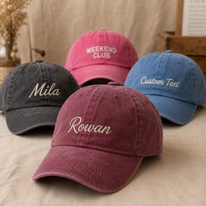 Könnte beinhalten: Vier Baseballkappen in Anthrazit, Pink, Bordeauxrot und Blau. Die anthrazitfarbene Kappe trägt den Namen Mila, die pinke Kappe zeigt WEEKEND CLUB, die bordeauxrote Kappe zeigt Rowan und die blaue Kappe zeigt Custom Text in weißer Stickerei.