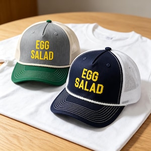 Könnte beinhalten: Zwei Baseballkappen mit dem Text "EGG SALAD" in Gelb. Eine Kappe ist marineblau mit weißem Mesh-Rücken und Schirm, die andere ist grau mit beigem Mesh-Rücken und grünem Schirm. Beide liegen auf einem weißen T-Shirt.
