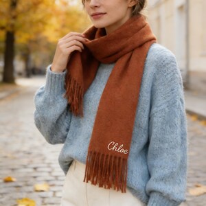 May include: A rust-colored scarf with fringe detail and the name "Chloe" embroidered in white script. The scarf is draped over a light blue sweater. The image is taken outdoors, with a blurred background of trees and a paved walkway.