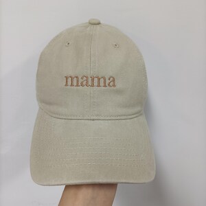May include: A beige baseball cap featuring the word mama embroidered in brown thread across the front center. The cap has a structured crown, a curved brim with visible stitching, and small ventilation eyelets on the top panels.