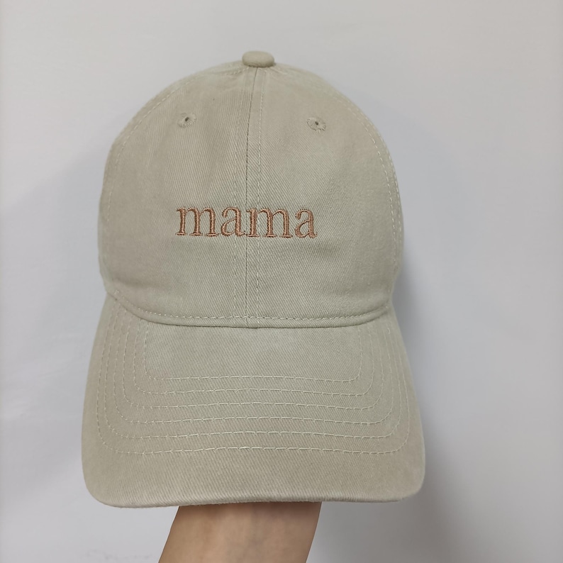 May include: A beige baseball cap featuring the word mama embroidered in brown thread across the front center. The cap has a structured crown, a curved brim with visible stitching, and small ventilation eyelets on the top panels.