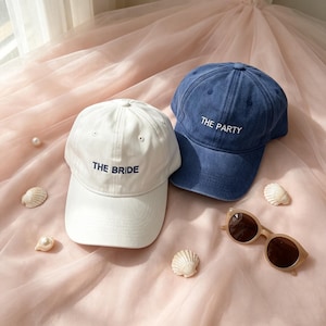 May include: A white baseball cap with the text THE BRIDE embroidered in dark blue and a blue baseball cap with the text THE PARTY embroidered in white, accompanied by a pair of brown-tinted sunglasses and several small white seashells.