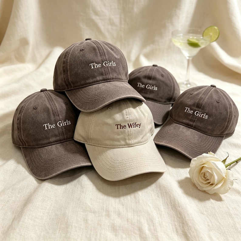 May include: A collection of four baseball caps in a distressed charcoal gray finish and one beige baseball cap. The charcoal caps feature the embroidered text The Girls in white, while the beige cap features the embroidered text The Wifey.