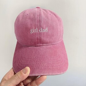 May include: A dusty rose pink baseball cap featuring the text girl dad embroidered in white thread on the front panel. The cap has a structured crown, a curved brim with visible stitching, and eyelets for ventilation on the top panels.