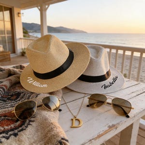 Könnte beinhalten: Zwei Fedora-Hüte, einer beige und einer grau, mit schwarzen Bändern und personalisierten Namen. Goldgerahmte Sonnenbrillen und eine goldene Anhänger-Halskette mit dem Buchstaben "D" liegen auf einem weißen Holztisch. Eine gemusterte Decke.