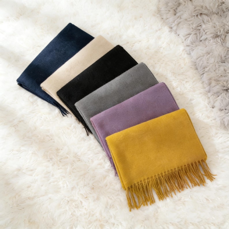May include: A collection of six rectangular scarves in various colors, including navy, beige, black, gray, purple, and mustard yellow. Each scarf has a fringed edge. The scarves are arranged diagonally on a white, fluffy surface.