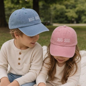 Könnte beinhalten: Eine blaue Baseballkappe mit dem weiß gestickten Text BIG BRO und eine rosafarbene Baseballkappe mit dem weiß gestickten Text BIG SIS. Beide Kappen haben eine strukturierte Krone, einen gebogenen Schirm und einen Stoffriemen hinten.