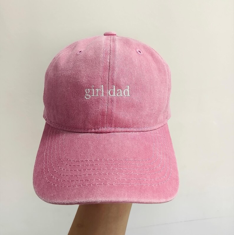 May include: A pink baseball cap with a washed, distressed texture. The front panel features the text "girl dad" embroidered in white, lowercase, serif font. The cap has a curved brim with visible stitching and a button at the top center.
