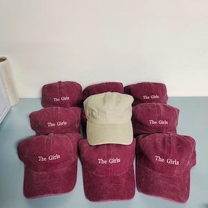 May include: A collection of baseball caps arranged in a group. Eight caps are deep maroon with the text The Girls embroidered in white, while one central cap is light beige with the text The Girls embroidered in a light pink thread color.