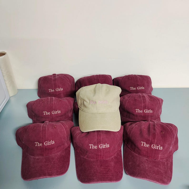 May include: A collection of baseball caps arranged in a group. Eight caps are deep maroon with the text The Girls embroidered in white, while one central cap is light beige with the text The Girls embroidered in a light pink thread color.