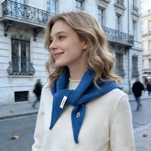 Könnte beinhalten: Eine Frau trägt einen blauen Schal und einen weißen Pullover. Der Schal hat ein kleines weißes Etikett und ein weiß gesticktes "S". Der Hintergrund zeigt eine europäische Straßenszene mit Gebäuden und Fußgängern.