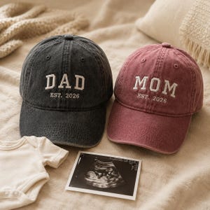 Könnte beinhalten: Zwei Baseballkappen, eine anthrazitgraue und eine altrosa, mit weißer Stickerei "DAD EST. 2026" und "MOM EST. 2026". Ein Ultraschallbild und ein Baby-Body sind ebenfalls im Bild.