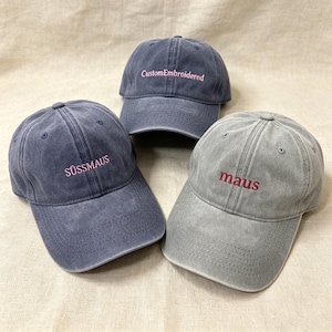 Peut inclure: Trois casquettes de baseball bleues et grises. Chaque casquette porte une inscription brodée dans une couleur contrastante. Une casquette indique "CustomEmbroidered", une autre "SÜSSMAUS" et la troisième "maus".