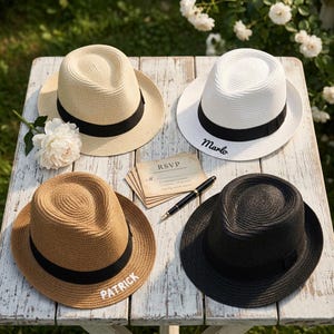 Könnte beinhalten: Vier Fedora-Hüte mit schwarzen Bändern liegen auf einem weißen Holztisch. Ein Hut ist beige, einer ist weiß mit gesticktem "Marlo", einer ist braun mit gesticktem "Patrick" und einer ist schwarz. Eine RSVP-Karte und ein Stift liegen ebenfalls auf dem Tisch.