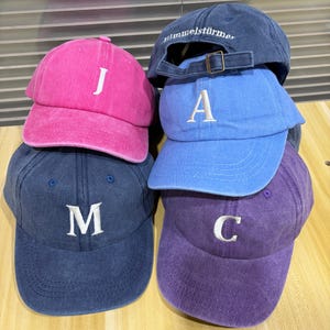 Könnte beinhalten: Eine Sammlung von vier Baseballkappen in Pink, Blau, Marineblau und Lila. Die pinke Kappe zeigt den Buchstaben J, die blaue den Buchstaben A, die marineblaue den Buchstaben M und die lila Kappe den Buchstaben C in weißer Stickerei.