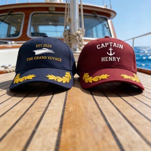 Könnte beinhalten: Zwei Baseballkappen auf einer Holzoberfläche. Eine ist marineblau mit goldenem Text "EST.2026" und "THE GRAND VOYAGE" sowie einem goldenen Blattdesign. Die andere ist weinrot mit "CAPTAIN HENRY" und einem Anker.