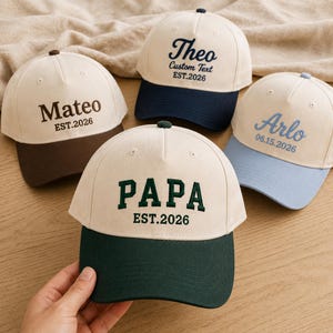 Puede incluir: Cuatro gorras de béisbol con texto personalizado. Las gorras tienen una corona color crema y una visera de color. El texto incluye nombres como "Mateo" y "Arlo", y la palabra "PAPA". Las gorras están bordadas con el año "EST.2026".