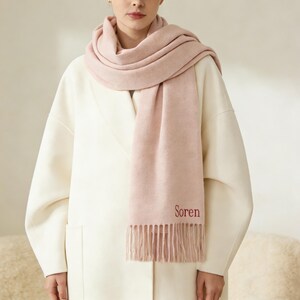 May include: A light pink scarf with the word "Soren" embroidered in red. The scarf is draped around the neck and shoulders, with a fringed edge. The person is wearing a white coat.