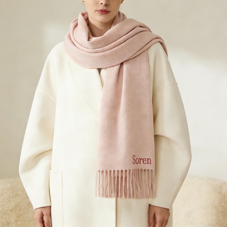 May include: A light pink scarf with the word "Soren" embroidered in red. The scarf is draped around the neck and shoulders, with a fringed edge. The person is wearing a white coat.