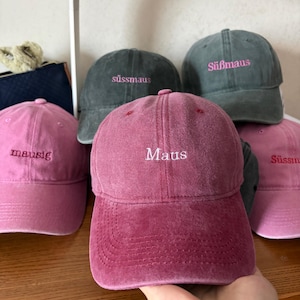 Könnte beinhalten: Eine Sammlung von sechs Baseballkappen in Rosa- und Dunkelgrautönen. Jede Kappe ist auf der Vorderseite mit Text bestickt, darunter die Wörter Maus, mausig und Süßmaus in weißem oder rosafarbenem Garn. Die Kappen haben eine weiche, verwaschene Stoffstruktur.