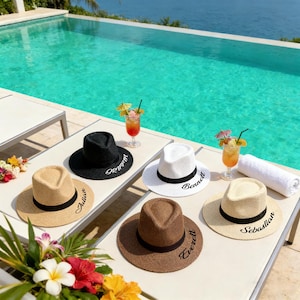 Könnte beinhalten: Eine Auswahl an Strohhüten in Schwarz, Weiß, Beige und Braun, jeweils mit einem schwarzen Band und einem Namen. Die Hüte liegen neben einem Pool mit Cocktails, was eine Urlaubsstimmung erzeugt.