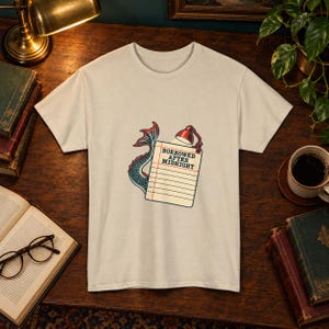 Può includere: Una t-shirt a maniche corte color crema con la grafica di un blocco note a righe illuminato da una lampada da scrivania rossa. Una coda di sirena blu e rossa si avvolge attorno al blocco note. Il testo BORROWED AFTER MIDNIGHT è stampato sopra.