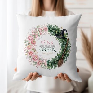 Pode incluir: Almofada quadrada branca com um design de grinalda floral. A grinalda é dividida em duas metades, uma com rosas cor-de-rosa e a outra com folhas verdes, um chapéu de bruxa e uma vassoura. O texto "PINK GOES GOOD WITH GREEN" está no centro.