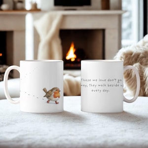 May include: Two white ceramic mugs. One mug features a watercolour illustration of a robin with a red breast and a trail of footprints. The other mug has the text: "Those we love don't go away, they walk beside us every day."