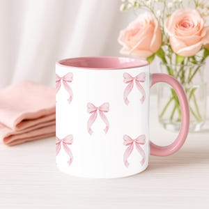 May include: White ceramic mug with a pink interior and handle. The exterior has a repeating pattern of watercolour-style pink bows. The mug is on a white surface, with a pink cloth and a vase of pink roses in the background.