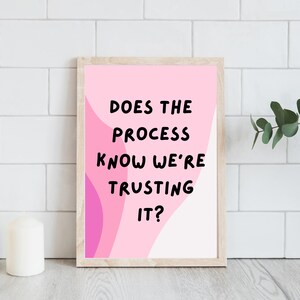 Op de afbeelding: Een ingelijste print met de tekst "DOES THE PROCESS KNOW WE'RE TRUSTING IT?" in zwart op een roze en witte achtergrond. De print zit in een licht houten lijst en wordt weergegeven op een wit oppervlak met twee witte kaarsen en groen.